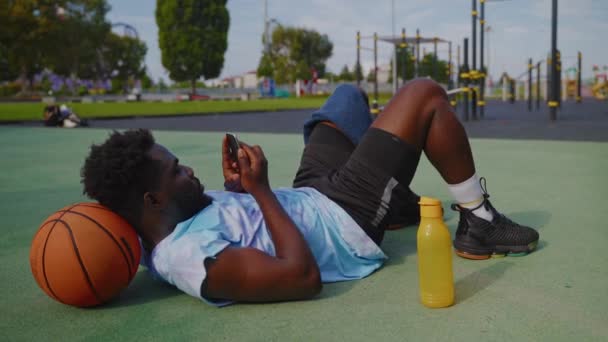 Afrikkalainen Amerikkalainen Kaveri Käyttää Älypuhelinta Makaa Kentällä Kadulla Koripallokenttä Urheilija — kuvapankkivideo