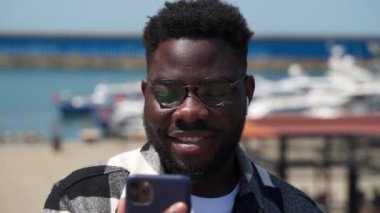 Elinde akıllı telefonla şehir limanında dikilen, modern aletlerle genç siyahi bir adamın gülümseyerek sosyal ağlarda komik videolar izleyen neşeli Afrikalı Amerikalı adamın portresi.