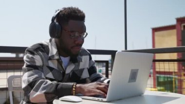 ses tasarımcısı veya müzik yapımcısı kulaklıkla müzik dinliyor, laptop ekranına bakıyor, sokak kafesinin açık terasında çalışan siyahi adamın portresi, Afrikalı Amerikalı serbest çalışan adam