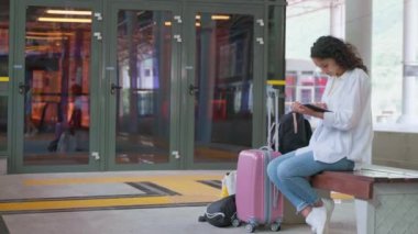 Tren istasyonunda trenini bekleyen bir sürü seyahat çantası olan genç bir kadın. Gitmeden önceki zamanı doldurmak için tablet bilgisayar kullanan kız