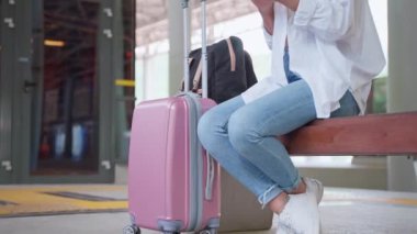 Bavullu genç esmer, platformda bankta oturuyor. Trenin kalkmasını beklerken akıllı telefon kullanan bir kadın gezgin.