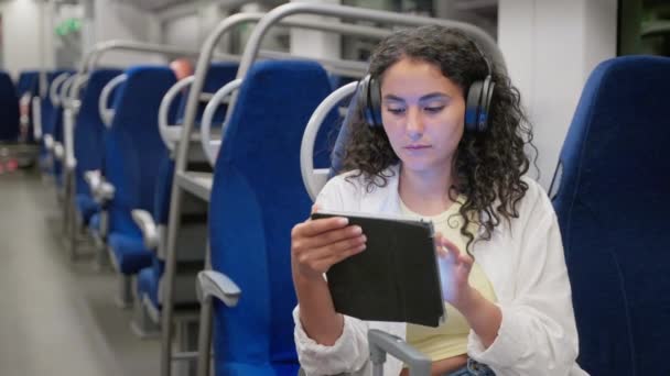 ガジェットと電車で旅行 ワイヤレスヘッドフォンで音楽を聴き タブレットコンピュータでネットサーフィンをする若い女性 旅行中の娯楽 — ストック動画