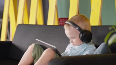 Küçük çocuk tablet bilgisayardan etkileniyor. Küçük çocuk evde vakit geçiriyor ve aletin üzerinde ilginç bir şey izliyor. Kablosuz kulaklık takan bir çocuk.