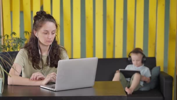 ラップトップを搭載したイヤホンの女性は 仕事やオンライントレーニングコースに集中しています 後ろのソファの上で小さな息子がタブレットPcで遊んでいる 仕事と育児を組み合わせたフリーランスの母親 — ストック動画