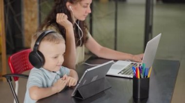 Kulaklıklı küçük çocuk, annesini rahatsız etmesin diye dijital tablette çizgi film izliyor. Evden çalışan bir kadın ve iş meselelerini çözmek için bilgisayarda video görüşmesi yapan bir kadın.