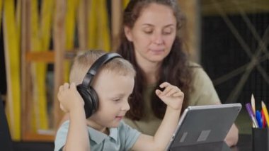 Anne, küçük oğlunun dijital tablet kullanarak yeni şeyler öğrenmesine yardım ediyor. Büyük kulaklıklı çocuk meraklı.