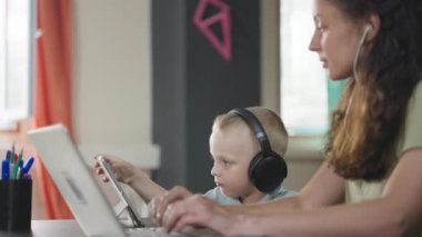 Kulaklıklı bir kadın dizüstü bilgisayar kullanıyor ve iş hakkında konuşuyor. Kulaklıklı küçük çocuk dijital tabletle oynuyor. Serbest meslek ve çocuk bakımı