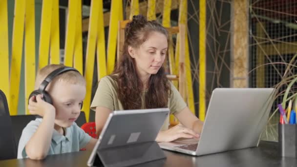 ヘッドフォンの小さな子供は 忙しい母親が家から遠く離れてラップトップを使用しているのを邪魔することなく デジタルタブレットで時間を楽しんでいます 家族と経歴 — ストック動画