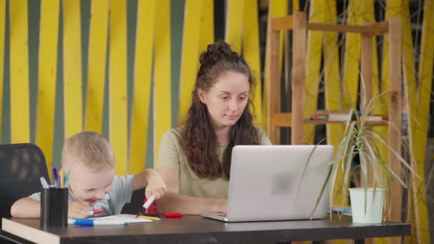 母親が仕事をしている間に遊んで絵を描く小さな男の子 リモートで働く女性とラップトップを使用しています フリーランスと家族 — ストック動画