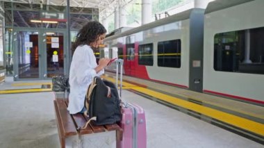 Genç bayan yolcu platformda trende bekliyor, bankta oturuyor ve terminalde ücretsiz kablosuz akıllı telefon kullanıyor, banliyöde trenle seyahat ediyor ve modern tren istasyonu