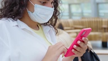 Yüz maskeli bayan yolcular internet üzerinden haberleri akıllı telefondan okuyor, bekleyen uçak veya tren, cihazla yüz ve ellerin kapatılması, terminalde ücretsiz kablosuz internet kullanımı, kalkış salonundaki kadın