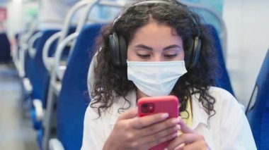Yüz maskeli genç hasta kadın trenle seyahat ediyor, toplu taşımada portre bayan yolcu, akıllı telefon ve kulaklık kullanıyor, internette ve sosyal medyada sörf yapıyor, Covid-19 salgını