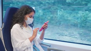 Trendeki Covid-19 virüs enfeksiyonundan korunmak için yüz maskesi takan kadın, seyahat ediyor ve akıllı telefon kullanıyor, internette sörf yapıyor, gazete okuyor, internet bağımlılığı bozukluğu