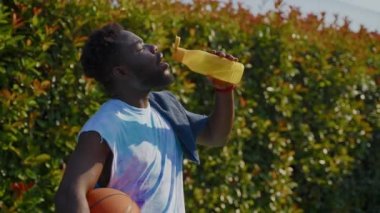 Siyah basketbolcu sıkı antrenmandan ya da açık sahada oynanan maçtan sonra plastik şişeden su içiyor, sokak sporu antrenmanı ve yarışması, Afrikalı Amerikalı sporcu susamış ve yavaş çekim hissediyor.