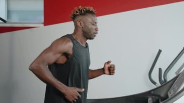 Siyah bir koşucunun yavaş çekim tarafı. Sporcu spor salonunda koşu bandında egzersiz yapıyor ve dijital ekrandaki egzersiz bilgilerine bakıyor. Kardiyo eğitimi