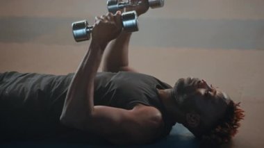 Siyah atletik bir adamın dikey dambıl kaldırışı, yatış pozisyonundan ağır çekimde. Sporcu silah gücü üzerinde çalışıyor.