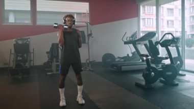 Atletik siyah bir adamın dambıllarla antrenman yaparken çekilen yavaş çekim çekimi. Sporcu biseps yapıyor ve kulaklıkla müzik dinliyor.