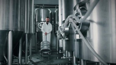 Bira fabrikası işçisi, büyük paslanmaz çelik bira işleme tanklarının arasında arabaya fıçıyı yuvarlıyor, ağır çekimde. Yiyecek ve içecek tesisleri