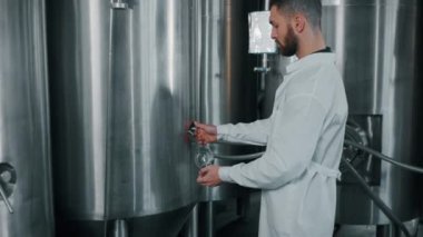 Profesyonel teknoloji uzmanı, bira fabrikasının atölyesindeki silindirik fermantasyon tankından, modern ekipmanlardan bira örneği alıyor.