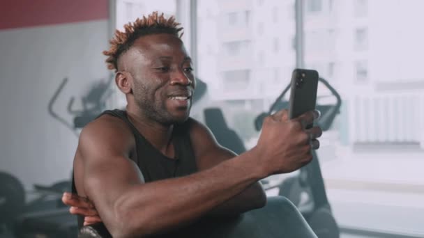 年轻的黑人运动员在健身房锻炼的过程中 正在社交网络上看有趣的视频 用智能手机描绘出一个微笑的非洲裔美国人的肖像 在健身中心使用手机 还有新的应用程序 — 图库视频影像