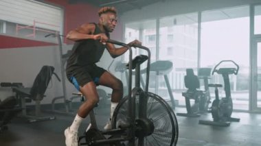 Yavaş çekim. Yorgun bir adam hava bisikleti üzerinde çalışırken elinden gelenin en iyisini yapıyor. Terli siyah sporcu tüm vücudunu çalıştırıyor.