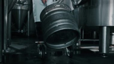 Bira fabrikasındaki paslanmaz çelik tankların arasında bira fıçısıyla el arabası yuvarlayan bir işçinin yavaş çekimi. İçecek üretimi
