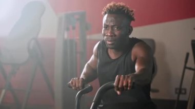 Sporcu Afrikalı Amerikalı adam spor salonunda egzersiz bisikletinin pedallarını çeviriyor, fitness merkezinde sıkı kardiyo eğitimi, formda kalmak ve kilo vermek için spor aktiviteleri, sağlıklı yaşam tarzı