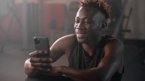 笑着的黑人男子在体育馆里用智能手机 锻炼后 用手机描绘出非洲共济会的形象 上网成瘾 男人用应用程序控制体重减轻和肌肉质量 — 图库视频影像