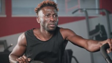 Şık kaslı adam spor salonunda antrenman yapıyor fitness merkezinde egzersiz makinesinde hareket ediyor, Afrikalı Amerikan sporcusunun portresi, sabit bisiklet üzerinde yoğun kardiyo çalışması, vücut geliştirme gücü eğitimi...