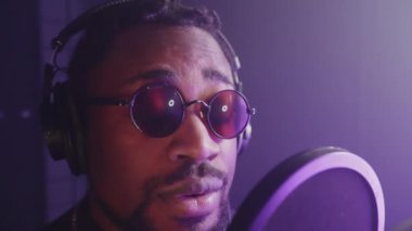 Gözlüklü siyahi bir şarkıcının mikrofona şarkı söylerken yavaş çekim görüntüsü. Ses geçirmez müzik stüdyosunda loş mor ışık
