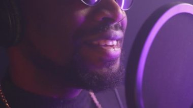 Mikrofona şarkı söyleyen sakallı zenci profesyonel bir müzisyenin yakın çekimi, yavaş çekim. Ses geçirmez stüdyoda müzik kaydı, loş mor ışıkta manzara