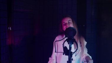 Genç ve enerjik bir kadının, müzik stüdyosunda mikrofona şarkı söylerken ve dans ederken yavaş çekim görüntüsü. Loş renk ışıklarıyla göster