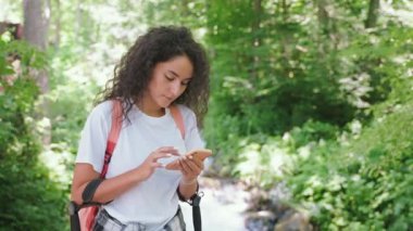 Ormanda nehirde yürüyüş yapan akıllı telefondan bakan kıvırcık bir kızın portresi. Dağ yürüyüşünde genç bir kadın Summer 'da haritada rota bulmak için telefondaydı. Turizm, seyahat, yürüyüş, gezi, macera konsepti.