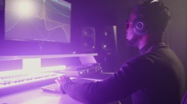 Müzik yapmak ya da karıştırmak için bilgisayar ve yazılım kullanan siyahi erkek ses üreticisi, kulaklıklardan keyif alıyor. Mor ışıklandırmalı ses stüdyosu