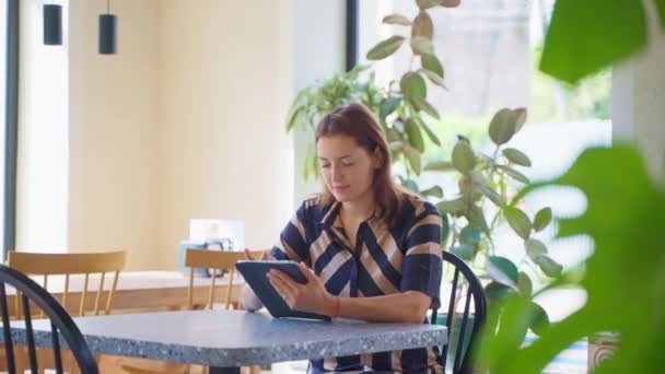 年轻而轻松的女人独自坐在咖啡店里 阅读信息 上网浏览平板电脑 女性休息等待命令 在社交媒体上聊天 观看内容滚动页面 做项目 — 图库视频影像