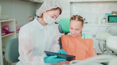 Modern stomatoloji kliniğinde çocuklar için ders, kadın dişçi ve genç kız tablet sergisine bakıyor, diş yapısını, hijyeni ve bakıcıların korunmasını konu alan videolar izliyor