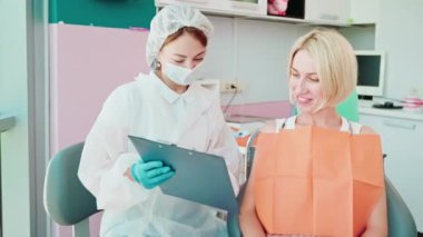 Diş hekimi, hastaya dişlerini nasıl tedavi edeceğini anlatıyor. Stomatolog doktor kadın klasörü elinde tutuyor ve gülümseyen genç bir kadına gösteriyor. Stomatoloji kliniği, dişçilik, sağlık kontrolü..