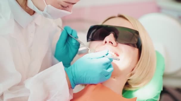 歯科鏡を用いた患者歯科医の歯科医学者で 症候診療所で歯科鏡を用いた歯科医の診察を行っています 歯科の黒い眼鏡のブロンドの若い女性は 歯のチェックアウトの予定 トップビュー — ストック動画