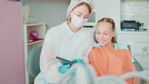 女性歯科医は 10代の少女の患者を笑顔にするためのタブレット上の教育ビデオを示しています 予防検査 歯科診療における歯科医による歯科診療に関する診察 — ストック動画