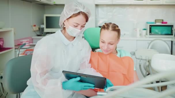 女性歯科医は 歯科の10代の少女患者へのタブレットのビデオを示しています 麻酔が発症するまで待っている少女との症候群学者は 症候群診療所で治療する前に効果を発揮します 歯科治療コンセプト — ストック動画
