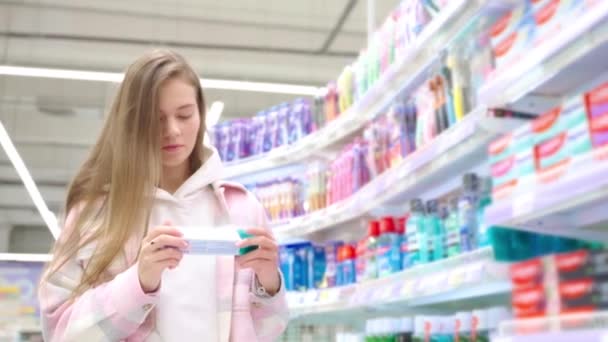 超级市场的年轻妇女在盒子里选择牙膏阅读配料 金发女孩站在展台附近购买带盒子和瓶子的牙科保健品 商店的购物理念 — 图库视频影像