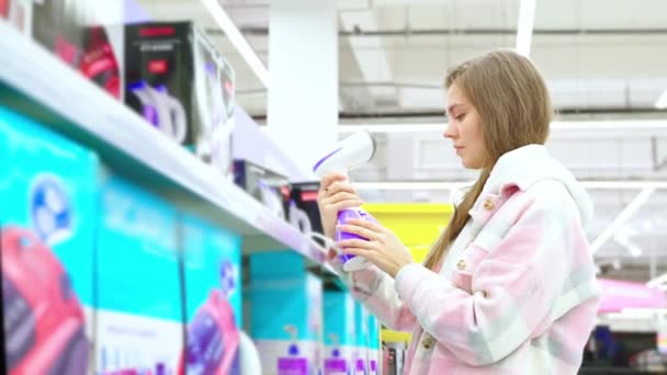 严肃的年轻女子选择在超市里买衣服蒸汽机 从橱窗 侧面看它 女孩在超级市场寻找家用电器 商店的购物理念 — 图库视频影像