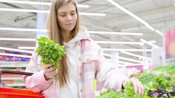 食料品店で緑の葉野菜を選択する若いブロンドの女性を買う スーパーマーケット ハイパーマーケットでグリーンを買う少女 パセリ グリーンオニオン ディル ビーガンエコ製品 健康食品コンセプト — ストック動画