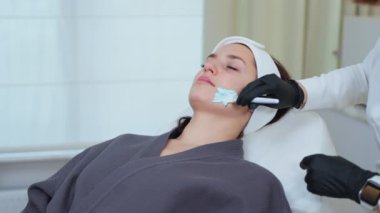 Spa salonunun kadın müşterisi cilt bakımı sırasında dinleniyor, kozmetolog kadın yüz derisi, anti-yaş ve sivilce güzellik prosedürlerine temizleyici ya da nemlendirici maske uyguluyor