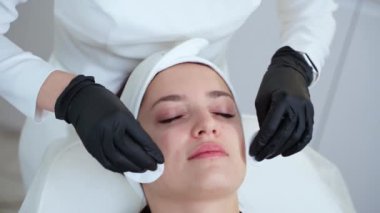 Yetişkin kadın cilt bakımı sırasında spa salonunda dinleniyor, estetikçi kadın hastaların yüzlerini nemlendirici losyonla siliyor, yüzüne yakın çekim yapıyor.
