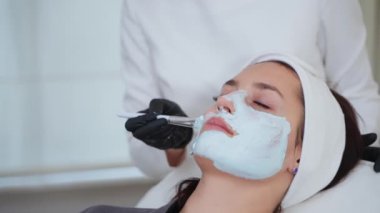 Estetikçi güzellik salonundaki kadın müşterinin cildine kilden yüz maskesi takıyor. Kozmetik kliniğinde rahatlatıcı ve gençleştirici bir tedavi uyguluyor.