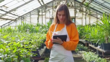 Bahçıvanlık ve bahçıvanlık, profesyonel çiçekçi kadın portresi serada büyüyen ev ve bahçe bitkilerini kontrol ediyor, bayan botanikçi notlar tutuyor.