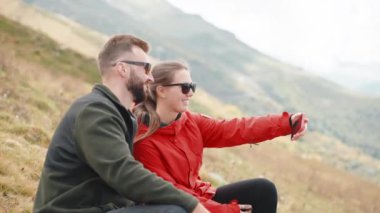 Turistler dağ yamacında otururken telefonla selfie çeken erkek yürüyüşçüler. Gezgin çift gezginler tatilde birlikte dağlarda ailelerini gezerler. İnsanlar kendilerini akıllı telefondan fotoğraflıyorlar..