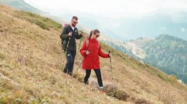 Genç adam ve kadın dağ yamacında yürüyorlar, yardım etmek için çubuklar kullanıyorlar, dağlık bölgede İskandinav yürüyüşü yapıyorlar, dağlarda turistler tatilde, bahar tatilinde aktif eğlence yapıyorlar.