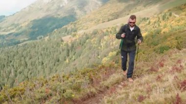 Sırt çantasıyla dağın tepesine tırmanan bir erkek yürüyüşçü. Kuzeyli yürüyüşü, dayanıklılığı ve doğada tek başına seyahat eden sportif bir adamın kuvveti için sopalar kullanıyor. Arka planda güzel dağlık bir arazi.